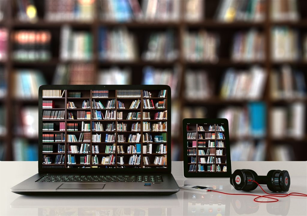 A table with an open laptop, a tablet, and a phone with headphones plugged into it. The laptop and tablet both show the same picture: a large bookshelf filled with books